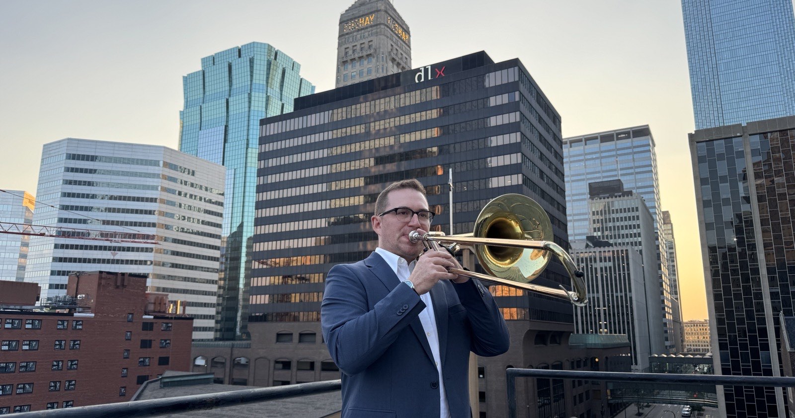 Christian Howard playing tenor trombone on a downtown Minneapolis rooftop, the city skyline rising behind him at sunset.